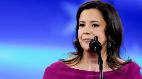 Representative Elise Stefanik stands before a microphone and in front of a blue backdrop. She wears a fuschia boat neck top. 