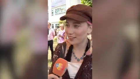 Young woman, wearing brown beret and hoop earrings, speaking into a microphone.