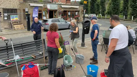 Carmel Wilson Five people are standing as they improve Ludlow. They are near a junction and buckets, bags and other pieces of cleaning equipment are on the ground and a bench.