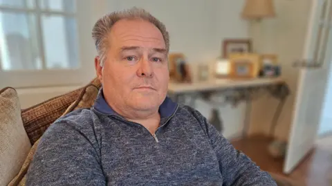 A man with short grey hair is looking at the camera. He is wearing a blue zip jumper and behind him is a table with photos.