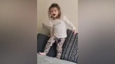 Cleveland Police Still from a video showing Isabelle running around on a grey sofa. She is smiling and excited. She is wearing flowery leggings and a white shirt and has shoulder length brown haair.