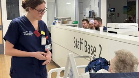 Charlotte Moore wears blue scrubs, glasses and has her brown hair tied back. She is wearing an NHS identification card as well as name badge and radio device. She is speaking to a grey-haired patient on a hospital bed. Two members of staff sit behind a reception area. 