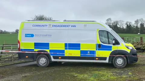 A community engagement police van. There is red police tape tied up in front of it and a field behind it with a red tent.