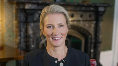 A blonde woman is smiling at the camera. Her hair is tied back and she is wearing a black round-necked jacket and silver pendant necklace. Behind her is an ornate fireplace.