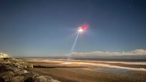 Paul Murphy/BBC Long exposure shot of a helicopter in flight over Withernsea. It is dark and there is bright light coming from the helicopter as it flies along the shoreline.