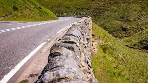 A stretch of the A57 Snake Pass showing a sudden drop at the side of a rocky wall. The wall is almost the same level as the road and there is a sharp bend towards the end of the road.