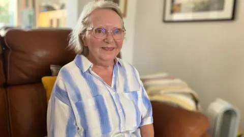 An elderly woman with white hair wearing glasses and a stripy blue shirt sits on a brown leather sofa.