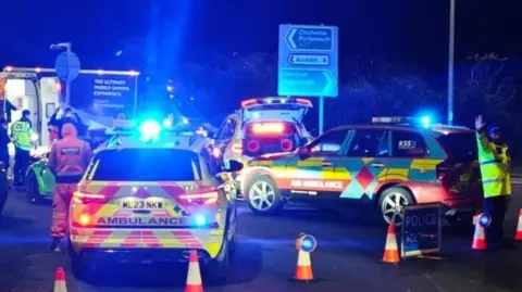 Eddie Mitchell Two police cars and an ambulance attend a scene at night. There are blue lights and traffic cones. 