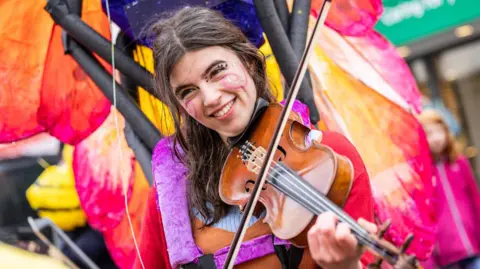 Paul Campbell A woman wearing pink and red clothing and face paints plays a fiddle. There are the colourful wings on a handcrafted insect behind her.
