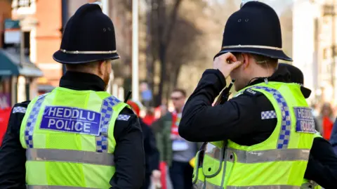 Two police officers wearing yellow high visibility jackets with "Heddlu" and "Police" written on the back.