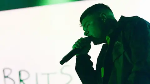 Móglaí Bap of Kneecap performs on stage at the O2 Academy in Glasgow. He is standing in front of a green and white background, with the word Brits written at the bottom corner.