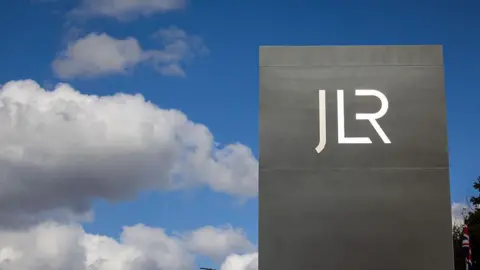 A large grey rectangular sign bearing the white JLR logo is photographed against a blue sky with scattered white clouds. A Union flag is partially visible behind the sign to the right. The image is taken from a low angle, making the sign appear prominent against the sky.