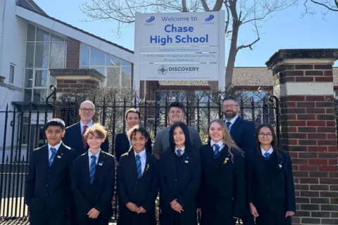 Discovery Educational Trust Ten staff and students stand for a group photo outside the Chase High School and one of its welcome signs. The children wear black blazers and blue striped ties. It is a sunny day.