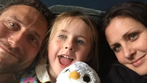 Steve Ackroyd, his daughter Autumn, and wife Fran smiling together on a couch, with Autumn holding a stuffed penguin toy.