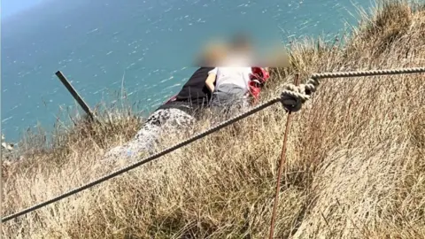 Eastbourne Coastguard Rescue Team Two people, their faces blurred, lie on the edge of the cliffs at Beachy Head looking over the edge, with the safety rope clearly closer to the camera.