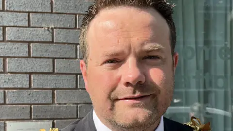 A man with short dark hair standing in front of a building. He wears a black suit, a white shirt and a red tie with a blue stripe on it. There is a gold chain hanging from his shoulders to represent his role as a mayor.