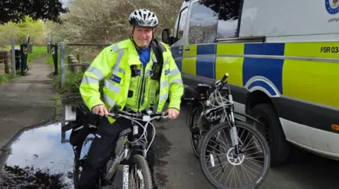 A police officer in a hi-vis jacket is on a bike and wearing a helmet. Trees and grass are in the background on the left and a police van is on the right.