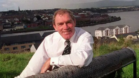 Ken Goodall, sits on a black park benc, in a grassy field. Behind him is a views of the city of Derry, including the river Foyle.