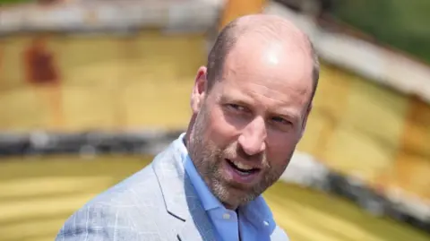 Prince William in a grey jacket and blue shirt. 