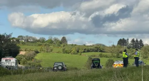 RTÉ Five vehicles parked in a field with two police officers standing to the right hand side of the image. There is a partially cloudy sky above.