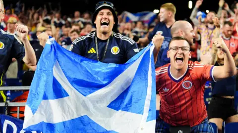 PA Media Scotland fans celebrate in the Boston stadium. In the foreground there is a man in a blue Scotland top and hat, holding a saltire flag. On his right is a man in a Scotland away strip with his left fist raised in celebration.