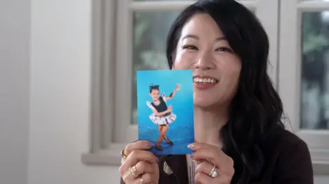 Picture of Arden Cho holding up a photograph of herself as a child. She has long, wavy, dark brown hair and is smiling at the camera. In the photograph she is at a dance contest, wearing a costume with a frilled skirt and sleeves, and is posing with her legs crossed and her hands held up in the air.