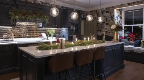 Kirsty Anderson A kitchen with cupboards made of navy wooden panelling. Christmas themed foliage, with lit candles in it, lies atop a large breakfast bar in the middle of the room. A garland  is also hung above the hob. 