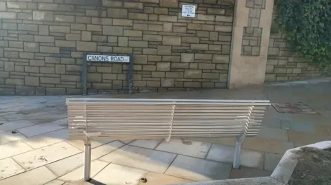 A metal bench facing a tan-coloured stone wall. The sign for Canons Road is in front of the wall, and a small 'no loading' sign can also be seen.
