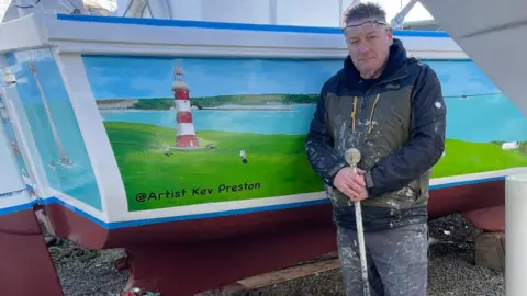 Kevin Preston Kevin Preston stands next to the boat. He has spikey brown hair and is wearing protective clothing covered in splashes of paint and is holding a white stick used while he paints. The mural has an image of a red and white lighthouse and 'Artist Kev Preston' written next to it.