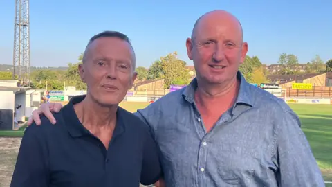 BBC Two men stand arm-in-arm on a sports ground in the sun.