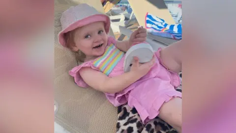 Sarah Normington A two-year-old child is lying on a sun lounger with a toy in her hand. She is wearing pink shorts and t-shirt and a matching hat.
