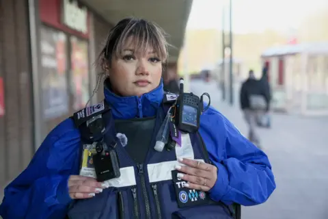 BBC/Andy Alcroft Esha Sheemar is wearing a blue jacket and protective vest. She is stood at a bus station, looking at the camera. 