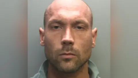 Samuel Hughes' mugshot. He has a shaved head and short stubble goatee.