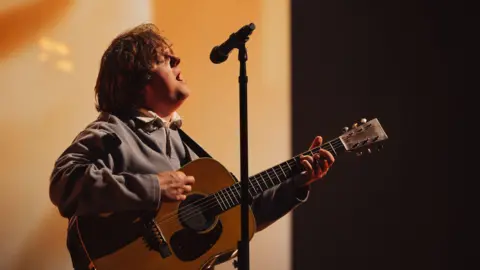 Lewis Capaldi sings into a microphone on a stand while playing an acoustic guitar. He has shoulder length hair and is wearing a grey sweatshirt.