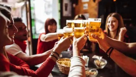 Getty Images Eight people near a table are holding their beer glasses together in this generic image. Four of them are smiling and some faces are not visible.