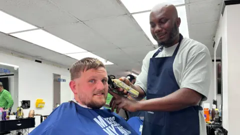 Emma Baugh/BBC Tom Clarkson is sitting down with a blue cape around him. A barber wearing a T-shirt and navy apron is shaving the side of his head.