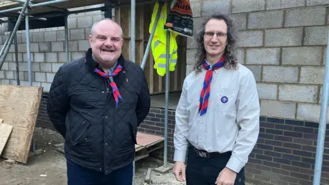 Amy Holmes/BBC A picture of two men, who are stood outside a new building that isn't finished yet. You can see the grey brick outer shell and scaffolding. The man on the left has short grey hair and is wearing a red and blue woggle and a black coat. The man on the right has long dark hair that is shoulder length and is also wearing a woggle but has a light grey shirt.
