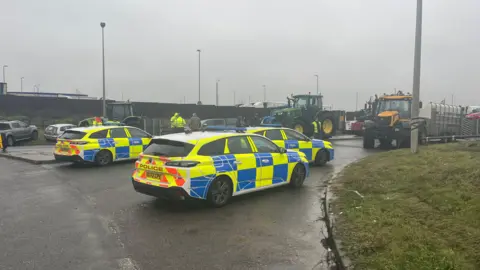 Tony Fisher/BBC Three police cars are parked in front of two tractors on a grey day