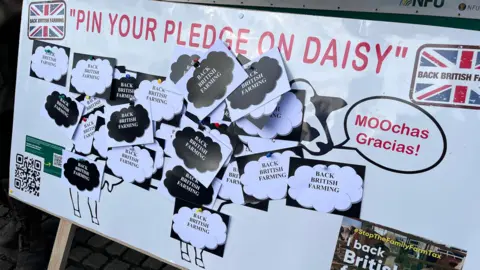 A white board with writing saying "pin your pledge on daisy". there are black and white pieces of card pinned to an image of a cow on the board. Each card says "Back British Farmers".