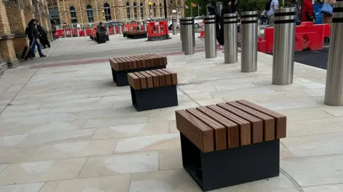 Flick Williams A line of small dark-coloured metal and wooden box seats. They are aligned at the opposite end with the tactile paving which leads across a Zebra crossing.