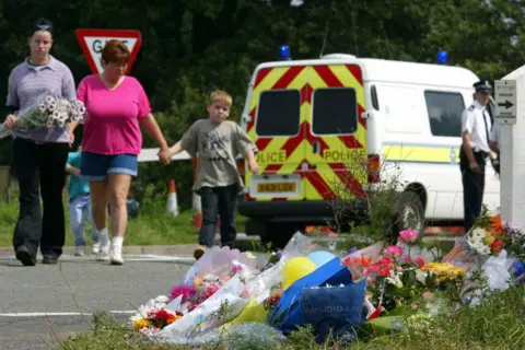 ODD ANDERSEN/AFP via Getty Images A woman in a pink T-shirt and blue denim shorts - and a boy in a sage green T-shirt - are let through a police cordon to lay flowers on a floral tribute. There are bunches of flowers on the grass in the foreground. There is a police van in the background. It is sunny.