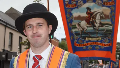 Pacemaker Archive photo: William Edgar from Ballykilbeg Orange Lodge attending last year's parade in Ballynahinch. He is wearing a black bowler hat, a white shirt, a red, white and blue tie, a blue suit jacket and an orange and purple sash, with gold fringes, around his shoulders. There is a large banner behind him depicting King William III on a white horse.