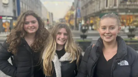 BBC Three women aged 18, one with long curly brown hair and black jacket, another with blonde hair and black jacket and another with blonde hair wearing black jacket with black top. They are in Belfast city centre.
