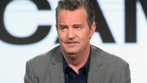 Actor Matthew Perry, wearing a grey suit jacket, speaking onstage at the 2017 Winter Television Critics Association Press Tour in Pasadena, California