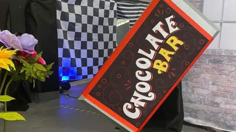Diagonally leaning sign reading “CHOCOLATE BAR” in white and yellow letters, set in front of black-and-white patterned backdrops and a brick-effect wall, with decorative flowers on the left.