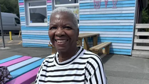 Hortense sat on a bench in front of a blue, white and pink ice cream parlour. She wears a black and white stripey t-shirt and has short blonde hair.