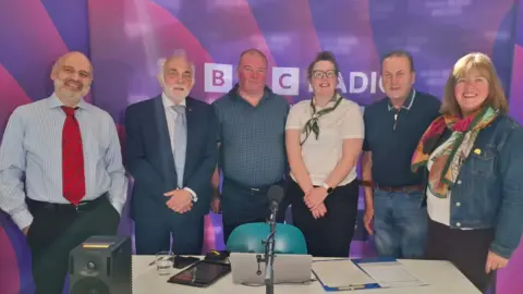Four men and two women stand in a row. They are standing in front of a large BBC Radio Devon purple banner. They are smartly dressed and are smiling at the camera. A microphone, laptop and speaker are on a table in front of them.