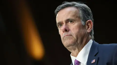 A suited John Ratcliffe is wearing a petrol blue suit and mauve tie with a USA flag on his left lapel. The pic is a head and shoulders shot with a microphone visible in front of him as if he is appearing at a hearing.