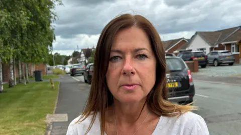 A woman who is wearing a white top is pictured standing in front of a road with cars and houses behind her. There are trees and grass on the left-hand side. 