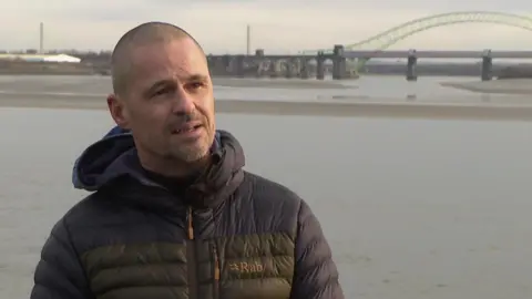 Ross has a shaved head and is wearing a black winter jacket. He is stood on the band of the River Mersey with a bridge behind him in the distance